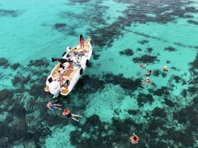 A lively group enjoys swimming in the crystal-clear turquoise waters, with a boat anchored nearby — perfect for a fun day of snorkeling and relaxation.