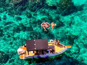 A lively group enjoys a fun day on the water, floating with colorful floaties while a boat is anchored nearby — perfect for summer adventures and group outings.