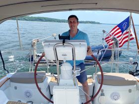 A man stands at the helm of a sailboat, ready to navigate the waters. The interior features a comfortable seating area and a clear view of the scenic surroundings.