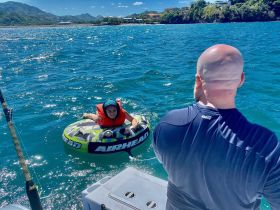 A young girl enjoys tubing in the clear blue waters, while an adult watches from the boat — a perfect moment of family fun on the water.