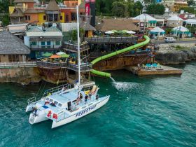 A lively scene featuring a catamaran named 'Freestyle' docked near a vibrant restaurant with a water slide, showcasing a fun atmosphere perfect for a day on the water.