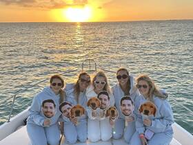 A group of friends celebrates at sunset on a boat, holding playful cutouts of faces — a perfect moment for fun and laughter on the water.