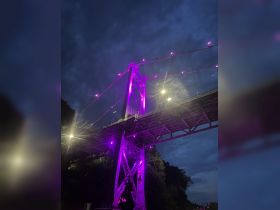 A nighttime view of a bridge illuminated with purple lights against a cloudy sky, creating a striking visual effect.