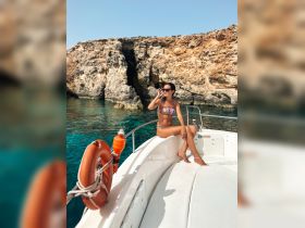 A woman enjoys the sun on the bow of a boat, surrounded by stunning rocky cliffs and crystal-clear waters — a perfect scene for a summer getaway.