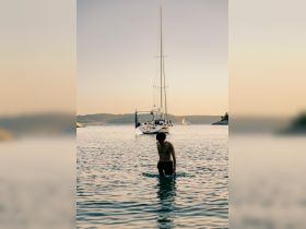 A person wades through calm waters near a sailboat at sunset, capturing the essence of a serene boating experience.