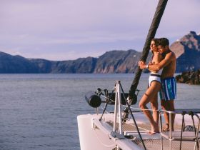 A couple enjoys a romantic moment on the bow of a boat, surrounded by stunning ocean views and mountains — perfect for memorable getaways.