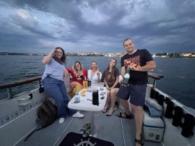 A lively group enjoys a boat party at dusk, celebrating with drinks and laughter against a scenic waterfront backdrop.