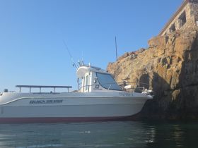A side view of the Quicksilver fishing boat 'NINA-1' docked near rocky terrain, showcasing its sturdy design against a clear blue sky.