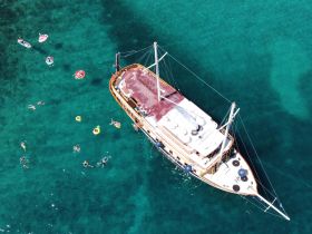 An aerial view captures a lively group enjoying the clear waters around a beautiful boat, with swimmers and inflatable tubes creating a vibrant scene perfect for summer fun.