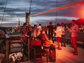 A lively group enjoys a festive evening on a boat, celebrating with cocktails and laughter against a stunning sunset backdrop.
