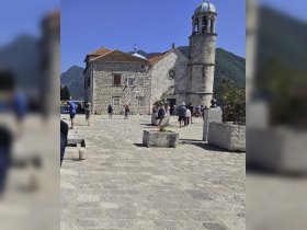 A bustling scene with people walking near a historic building, set against a mountainous backdrop. The image lacks clarity, making it difficult to discern specific details.