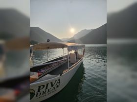 The Piva Dream boat is docked on a serene lake, framed by majestic mountains and a beautiful sunset, perfect for scenic cruises.