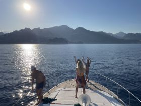 A lively group enjoys a sunset celebration on the deck of a yacht, dancing and soaking in the stunning views of the mountains and shimmering water.