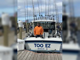 A smiling fisherman stands proudly on the deck of the 'TOO EZ' fishing boat in Muskegon, showcasing an array of fishing gear — perfect for a day out on the water.