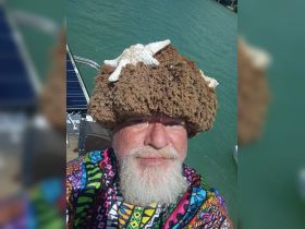 A man wearing a unique hat made of coral and starfish poses for a selfie on a boat, showcasing a vibrant outfit against a backdrop of turquoise waters.