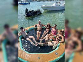 A lively group enjoys a fun day on a floating mat in the water, sipping drinks and soaking up the sun — perfect for summer parties and relaxation.