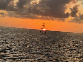 A stunning sunset over the ocean, with the sun setting behind a lighthouse structure, creating a picturesque scene perfect for evening boat trips.