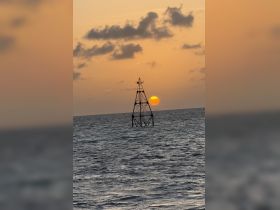 A silhouette of a structure against a sunset over the ocean, creating a serene yet ambiguous scene.