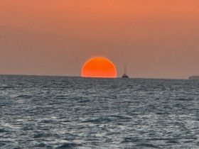 A stunning sunset casts a warm glow over the horizon, with a sailboat silhouetted against the vibrant sky — a perfect scene for tranquil evening cruises.