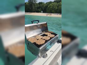 A grill loaded with patties on a boat, with a beautiful beach scene in the background — perfect for a fun day of barbecuing on the water.