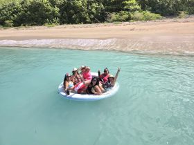 A lively group enjoys a fun day on a floating mat in crystal-clear waters, perfect for beach parties and relaxation under the sun.