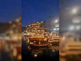 A boat is docked at a marina at night, with people visible on board, creating a lively atmosphere under the twinkling lights of the surrounding hillside.