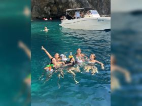 A lively group enjoys a fun day swimming in crystal-clear waters, surrounded by a boat and playful floaties — perfect for summer adventures.