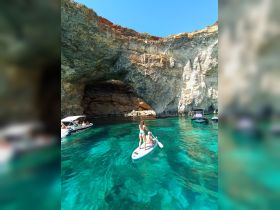 A couple enjoys paddleboarding in crystal-clear waters near a stunning cave, surrounded by boats — a perfect adventure in nature.