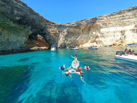 A lively group enjoys a sunny day swimming in crystal-clear waters near a stunning cave, surrounded by boats — a perfect summer adventure.