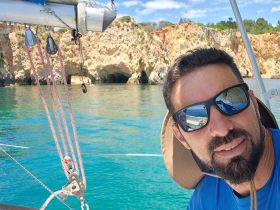 A sailor takes a selfie aboard a boat, showcasing the stunning ocean backdrop and rocky cliffs — a perfect moment of adventure on the water.
