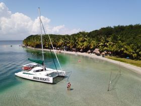 A couple enjoys a picturesque moment at the beach with a catamaran anchored nearby, surrounded by lush palm trees and clear waters — a perfect tropical getaway.