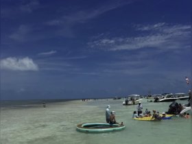 A vibrant sandbar scene with groups enjoying the water, lounging on inflatable mats, and surrounded by boats — perfect for a fun day out on the water.