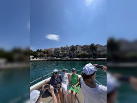 A group of friends enjoys a sunny day on a boat, wearing captain hats and capturing memories against a picturesque waterfront backdrop.