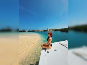 A young woman enjoys the sun while sitting on the edge of a boat, overlooking a beautiful beach and clear waters — a perfect moment for relaxation and fun on the water.