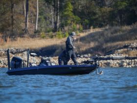 An angler stands on a fishing boat, reeling in a catch on a serene lake — a perfect moment for fishing enthusiasts.