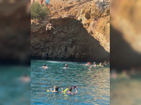 A thrilling moment captured as a child prepares to jump off a cliff while others enjoy swimming and snorkeling in the clear waters — a perfect highlight of a fun day on the water.