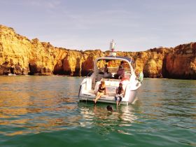 A lively group enjoys a sunny day on the water, with some swimming near the boat against a stunning coastal backdrop.