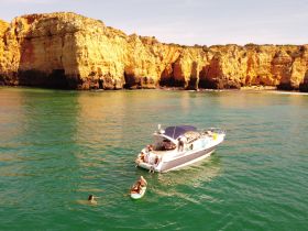 A vibrant scene of a group enjoying a sunny day on the water, with one person paddleboarding and others swimming near a boat, set against stunning coastal cliffs.