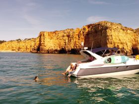 A lively group enjoys a sunny day on the water, with one person swimming near the boat and stunning coastal cliffs in the background — perfect for summer adventures.