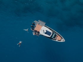 Aerial view of a group enjoying a refreshing swim around a boat in crystal-clear waters, showcasing the perfect summer day on the water.