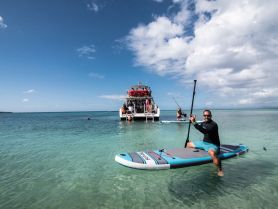A paddleboarder enjoys the clear waters while a group of guests relax on a boat in the background, capturing the essence of a fun day on the water.