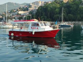A vibrant red boat named 'Vega' glides through calm waters, surrounded by picturesque hills and a charming coastal town.