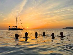 A group of friends enjoys a sunset swim in the ocean, with a beautiful yacht silhouetted against the vibrant sky — a perfect moment for relaxation and fun on the water.