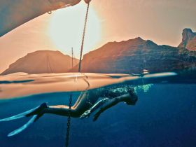 A stunning underwater shot of a snorkeler gliding through crystal-clear waters at sunset, with majestic mountains and boats in the background — a perfect representation of adventure and natural beauty.
