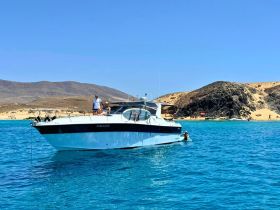 A couple enjoys a sunny day on the water, with one person swimming near the boat and a beautiful beach in the background — perfect for a relaxing getaway.