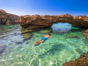 A snorkeler explores the crystal-clear waters beneath a natural rock arch, showcasing the beauty of underwater adventures in a stunning coastal setting.