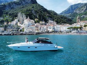 A joyful group aboard a sleek boat waves at the camera against a stunning coastal backdrop, showcasing the beauty of the Amalfi Coast.