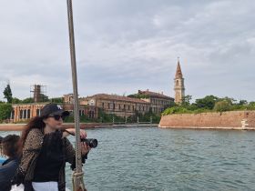 A woman captures the stunning scenery while aboard a boat, with historic architecture and a bell tower in the background, showcasing a picturesque moment on the water.