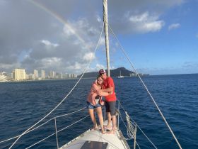 A couple enjoys a romantic moment on the bow of a sailboat, framed by a vibrant rainbow and the stunning skyline of Honolulu in the background.
