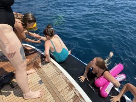 A lively group enjoys a fun day on the water, assisting each other as they swim and play off the side of a boat — perfect for summer adventures and bonding experiences.